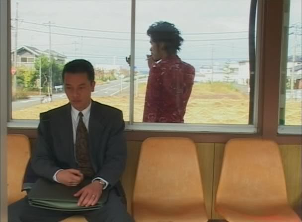 Miike, Takashi. Visitor Q. 2001. Man smoking behind another man who is seated, waiting at a bus stop