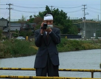 Miike, Takashi. Visitor Q. 2001. Man recording with a camcorder outdoors.