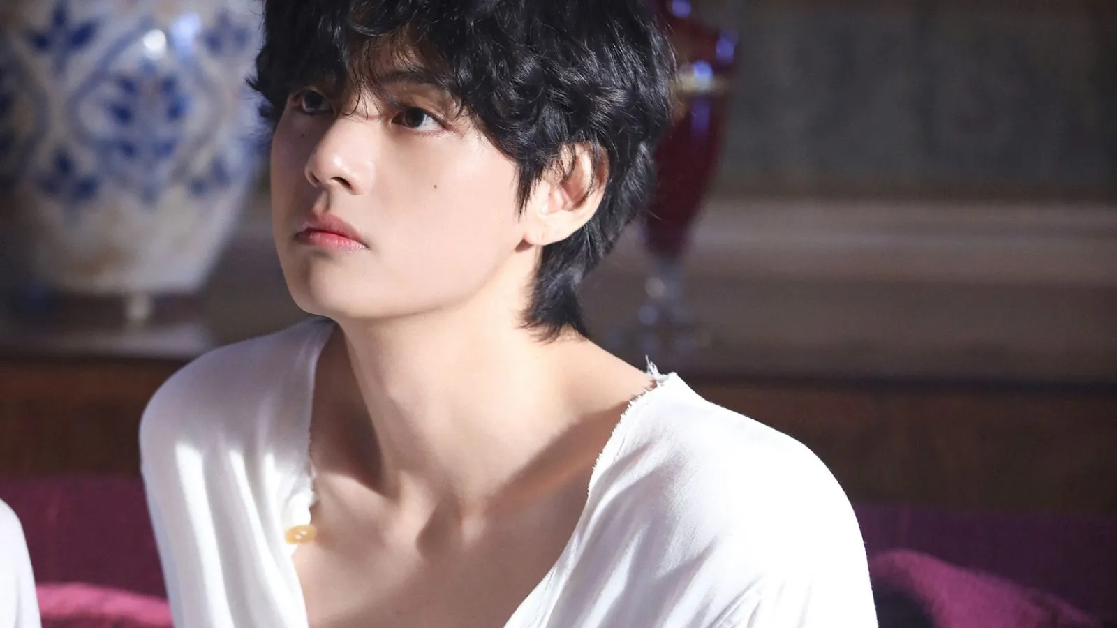 BTS member V looks to the side while wearing a white shirt.