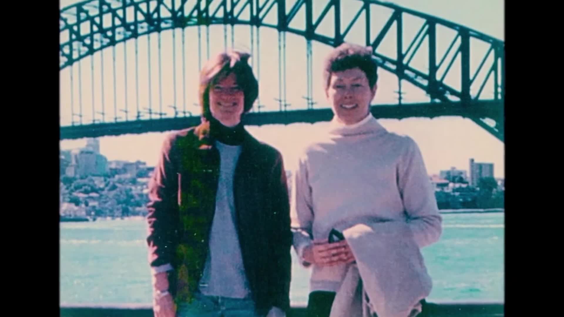 Sally Ride stands next to her life partner Tam O'Shaughnessy in front of a bridge. They are both looking at the camera and smiling.