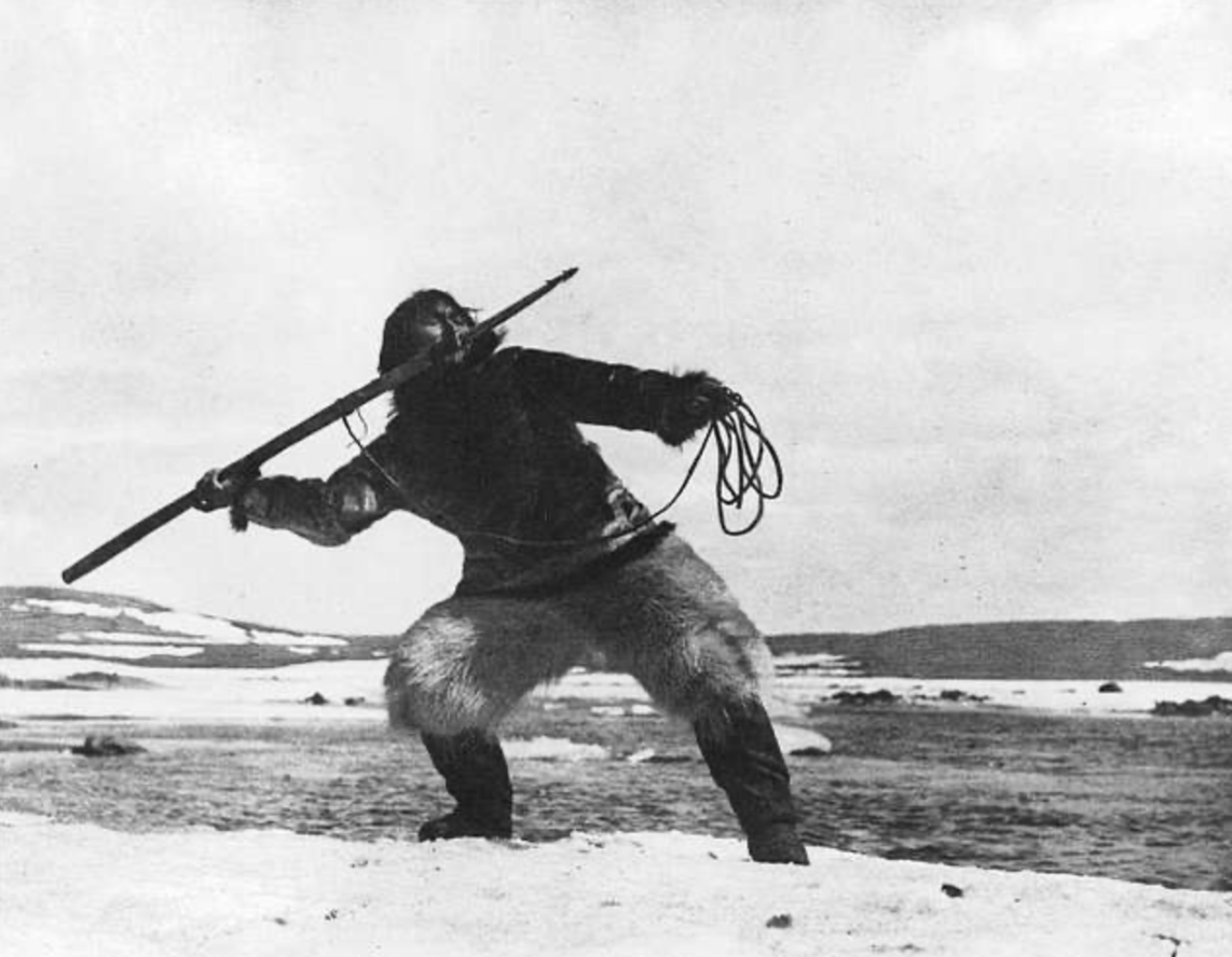 Allakariallak, renamed Nanook, holding a harpoon ready to throw and rope in his other hand, demonstrating traditional Inuit hunting techniques. He is standing on the snowy bank of an icy river.