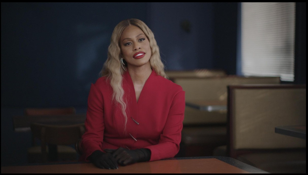 Laverne Cox sits in front of a camera alone in a dining room. She is wearing a red jacket that matches her red lipstick and black gloves.