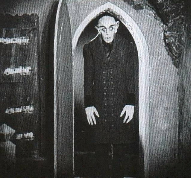 Nosferatu standing in the entryway of door that comes to a point at the top. He is almost as tall as the entrance. 