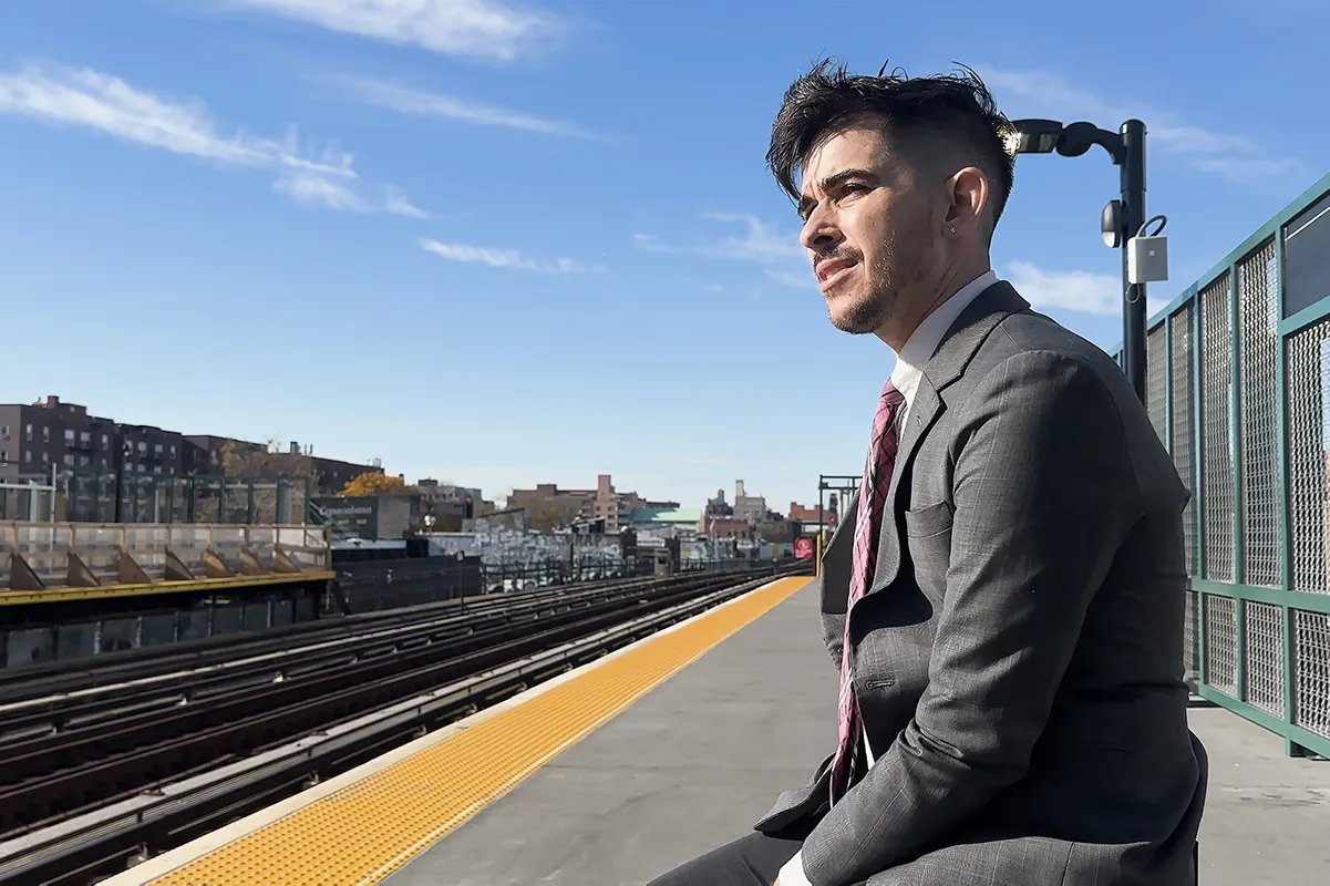 Chase Strangio sits on a bench at a train station. He is wearing a gray suit and red striped tie with the sun shining harshly on his face. The train station is otherwise empty.