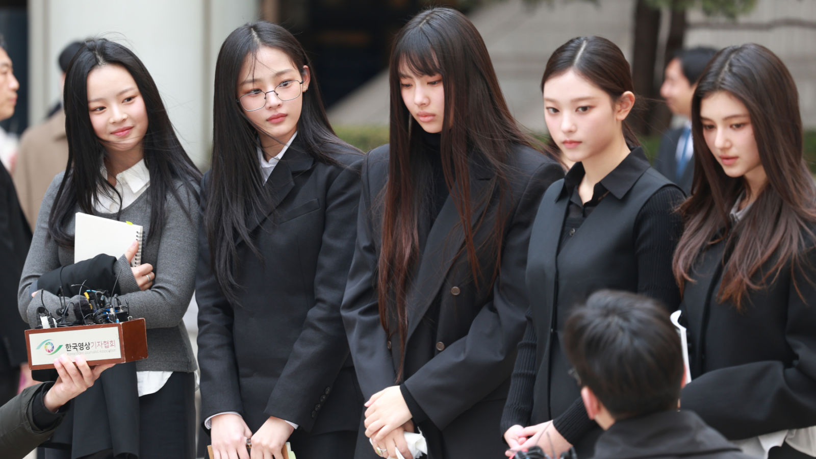 The members of NewJeans outside a court appearance, all dressed in dark neutral colors.