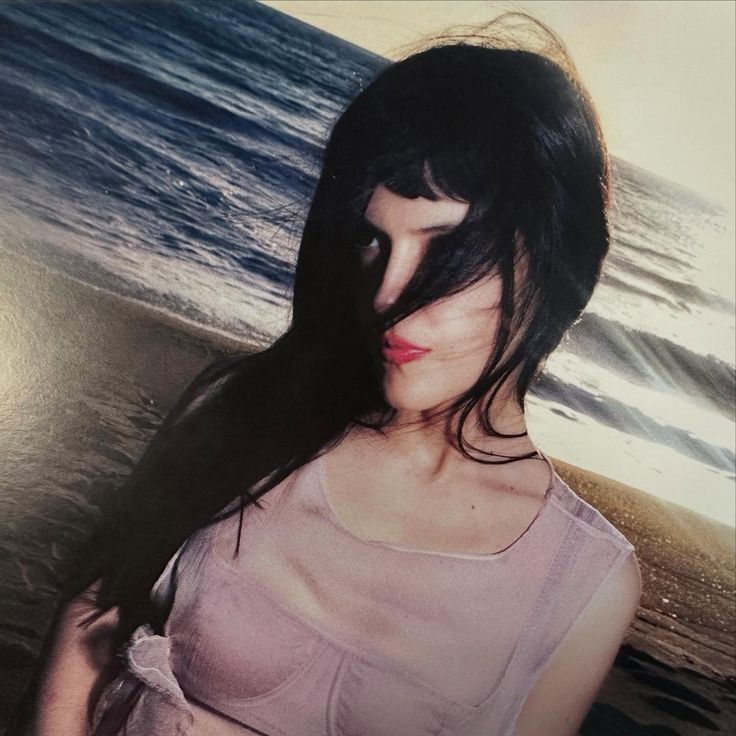 Maria Zardoya from The Marias is posing for a photo. There is an ocean and sand from the beach behind her. A strand of hair covers her right eye. The image is edited in Y2K style.