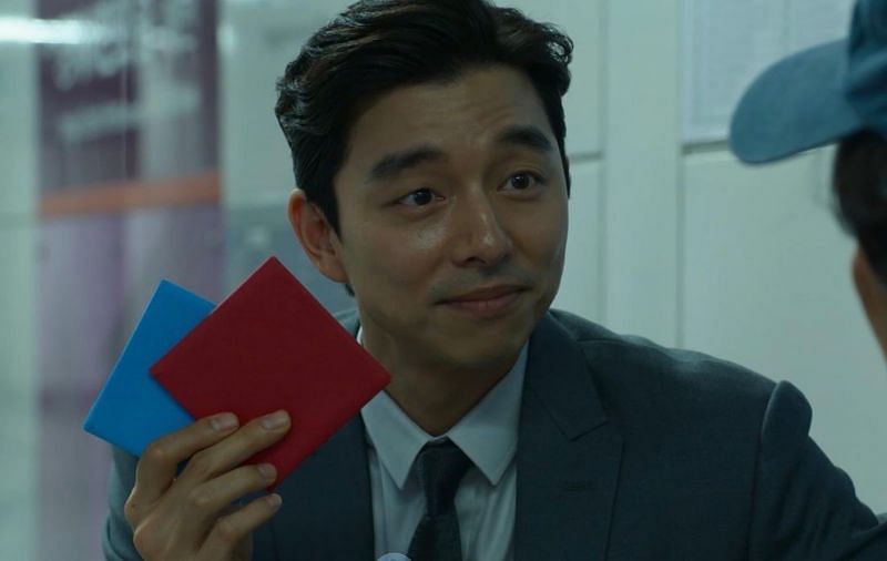 The recruiter holding the red and blue folded papers. He is wearing a suit and tie while the side of Gi-hun's face and blue hat can be seen. 