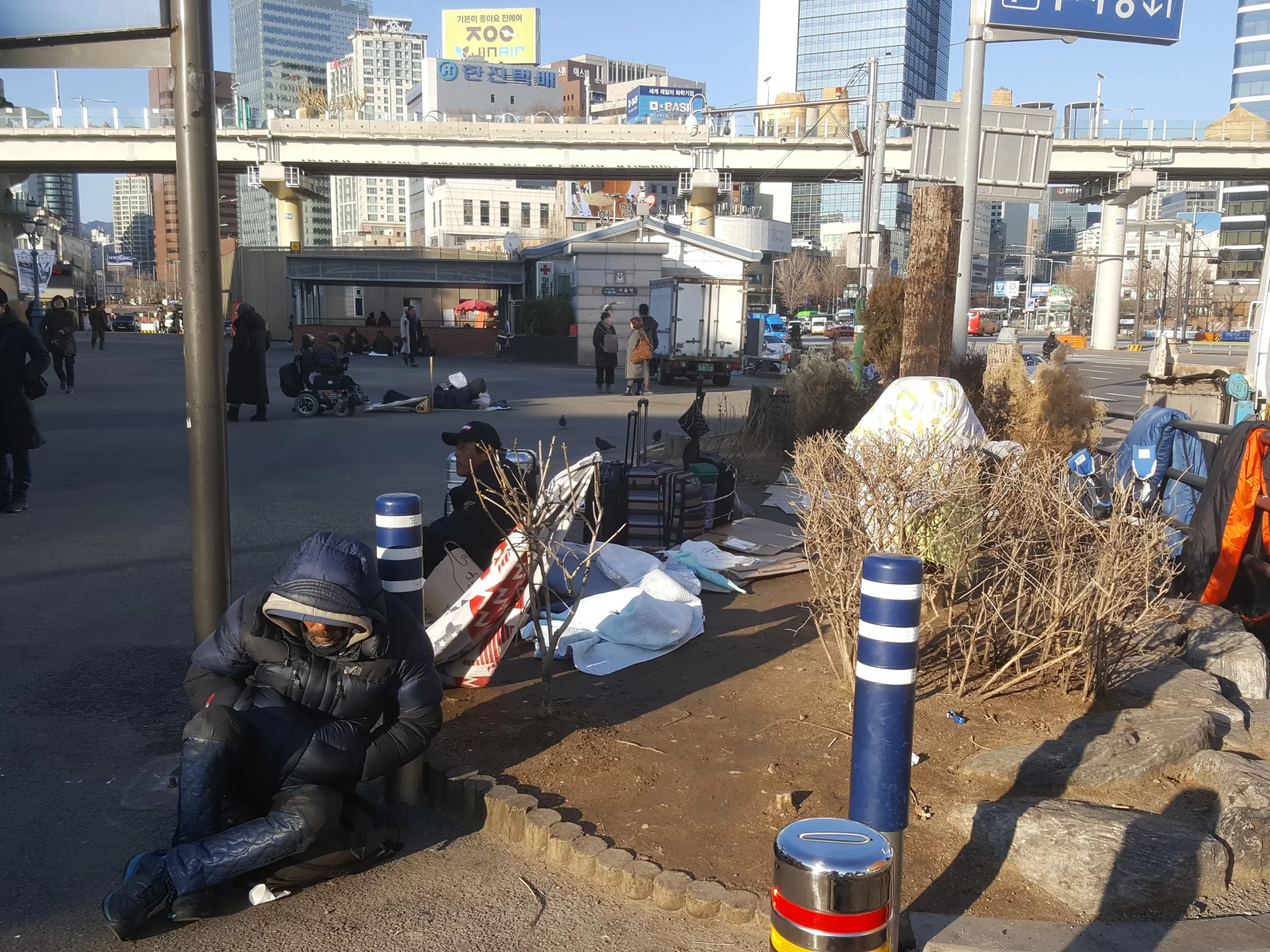 This image depicts the outside of Seoul Station at the corner of a block of metropolitan buildings. There are many roads in the background, but the foreground focuses on a dirt landscaping corner across from the Seoul station where homeless people have made makeshift camps with sleeping bags, suit cases, and cardboard. 
