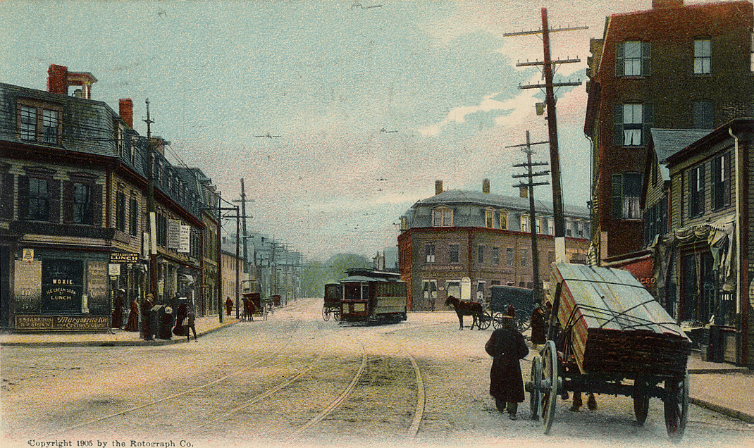 A dirt road lined by a couple of multi-storied buildings in Brookline, Massachusetts. There is a tram as welll as several horse-drawn carriages.