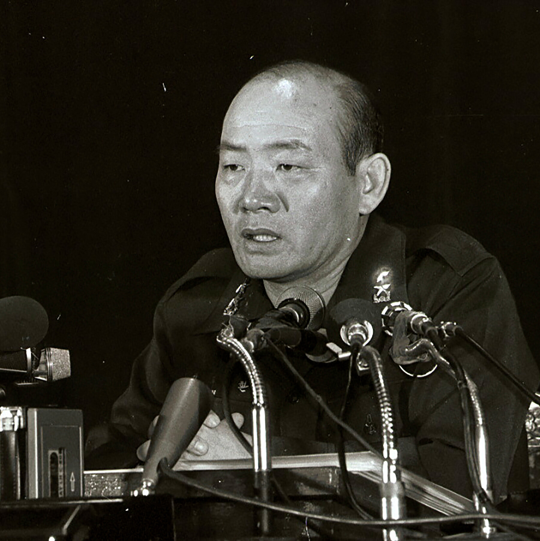 Chun Doo-hwan giving a press conference on October 28, 1979, two days after the assassination of Park Chung Hee. He is wearing a military uniform and speaking into multiple microphones.