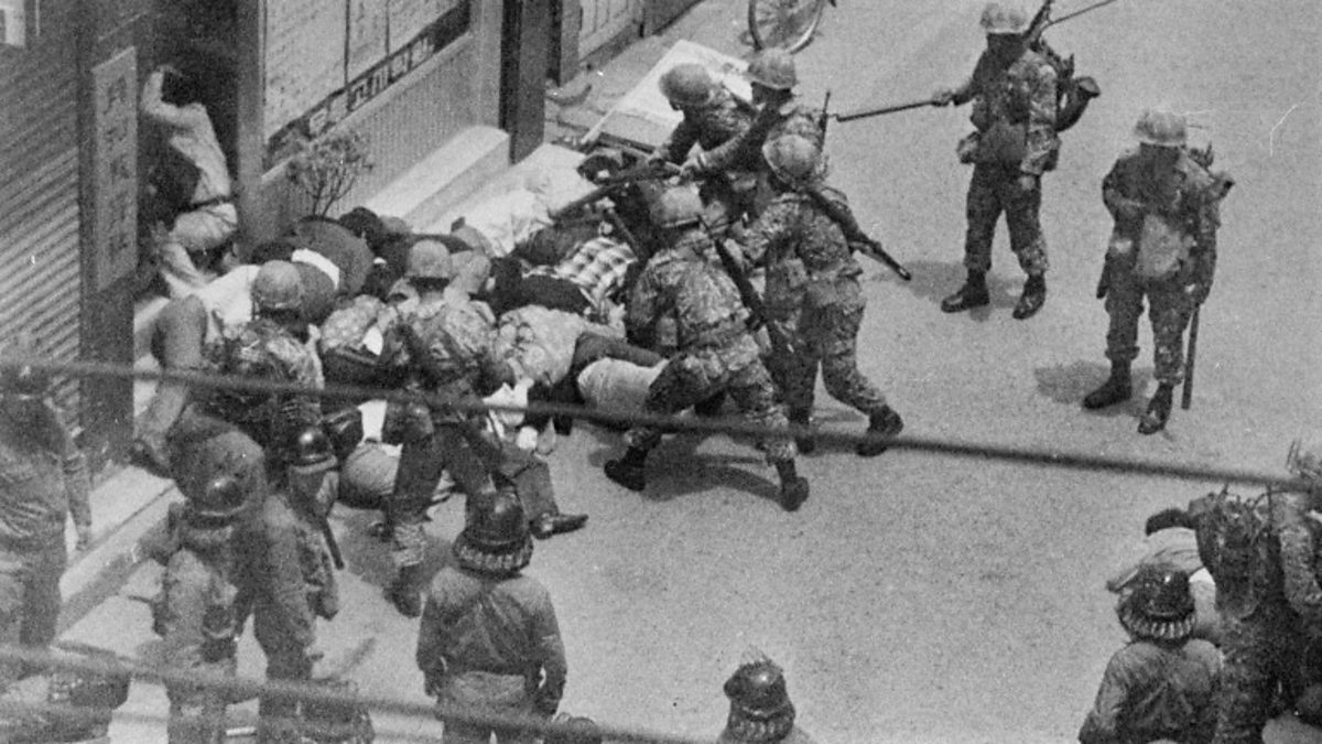 Military forces wielding batons over a crowd of plainly dressed people huddled in a pile at the doorsteps of a building. The image is from above, looking down onto the street where the clash is happening.