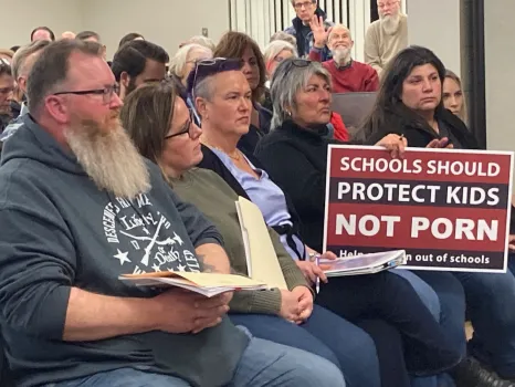 A group of Macob County, Michigan residents attending a school board meeting, one woman holding a sign that says "Schools should protect kids, not porn. Help...out of schools." The rest is covered by a woman's binder.