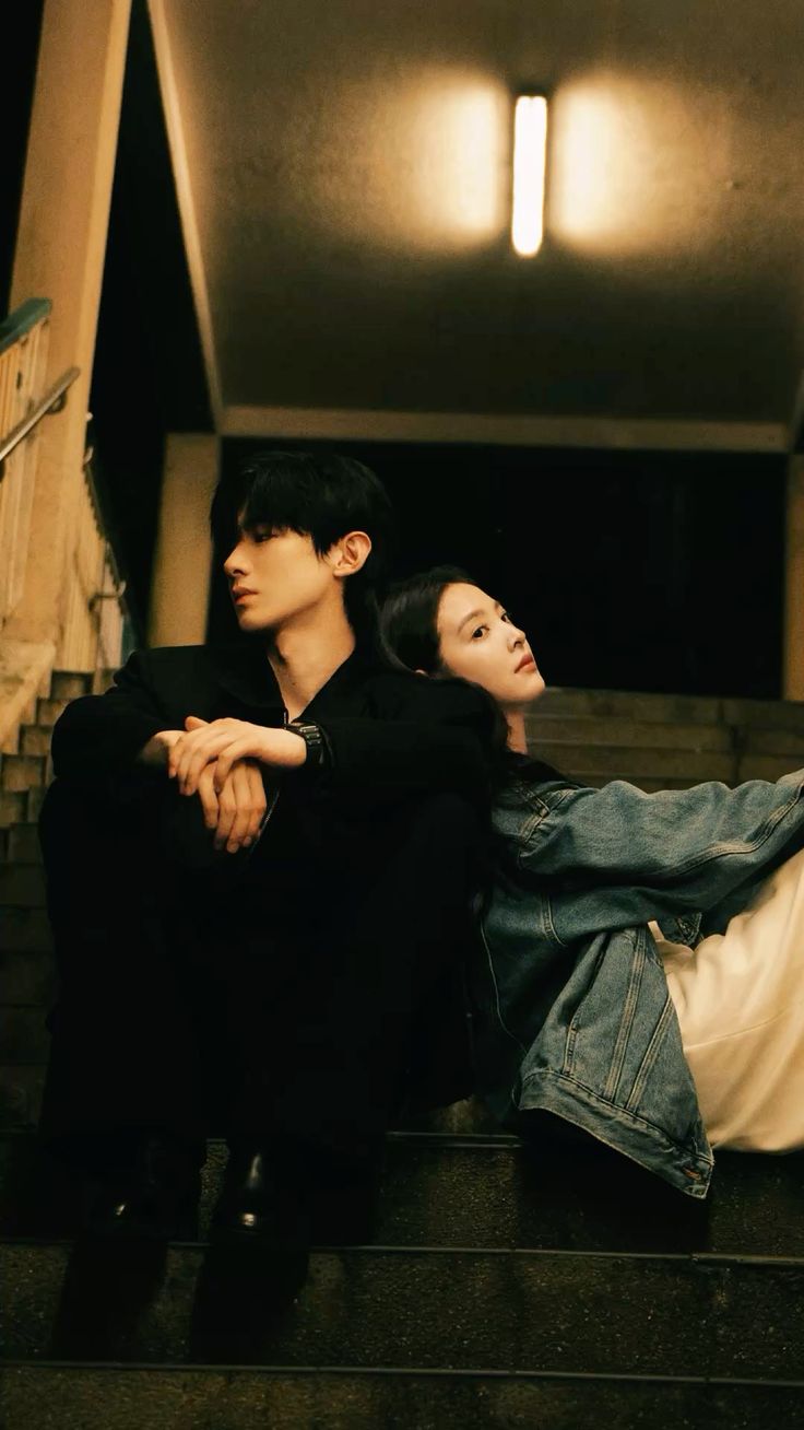 A man dressed in a black suit sits on a staircase with dim lighting and looks off to the left. A woman wearing a denim jacket and white pants sits next to the man, laying her head on his shoulder and looking upward.