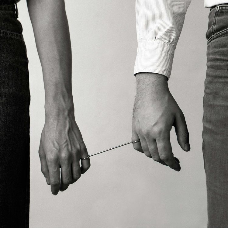 Two hands, the left is female presenting, and the right male presenting, are connected through rings. The rings are tethered together by a small rod, keeping the hands a certain distance away, but still close by. 