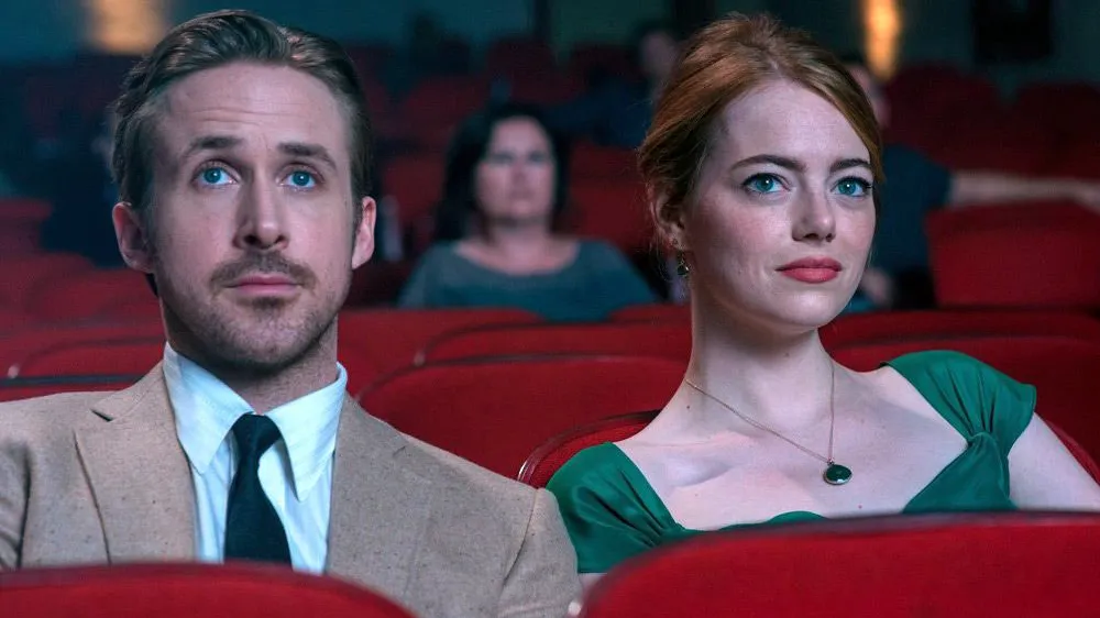 Seb and Mia are sitting next to each other in the theatre, staring at the screen ahead of them. Seb is in a tan suit while Mia is wearing a green dress, allowing them both to pop against the red seats.