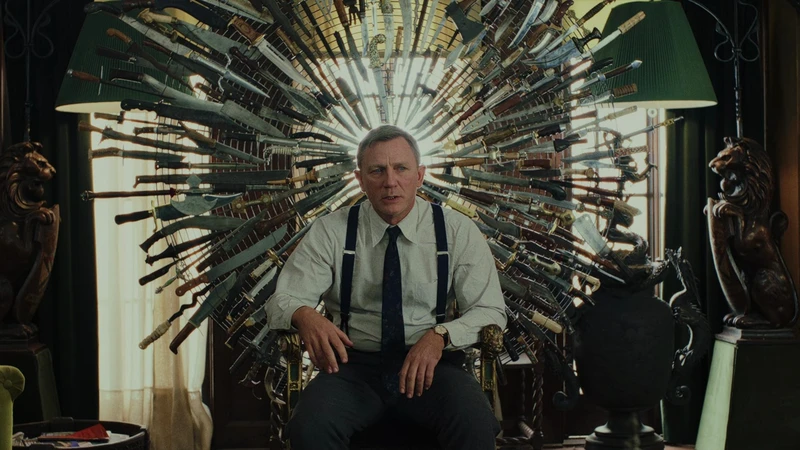 Benoit Blanc, played by Daniel Craig, sits slouched in a chair with a backing made into a circle of daggers pointing towards the center. He stares ahead of him, trying to figure out who the killer is. He's wearing a white button-up, black tie, suspenders, and trousers. The space around him in dark except for the covered window he's sitting in front of.