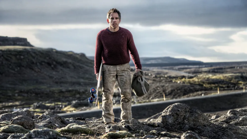 New Resolutions. Walter Mitty, played by Ben Stiller, stands in a rocky environment. The shot captures his full body as he stands carrying a skateboard and a bag. His appearance is disheveled, having traveled a long way to get to that point.