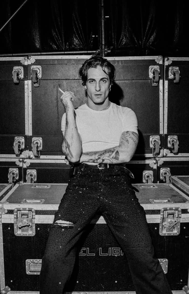 Damiano David is sitting on a compartment holding a cigarette between his fingers. 