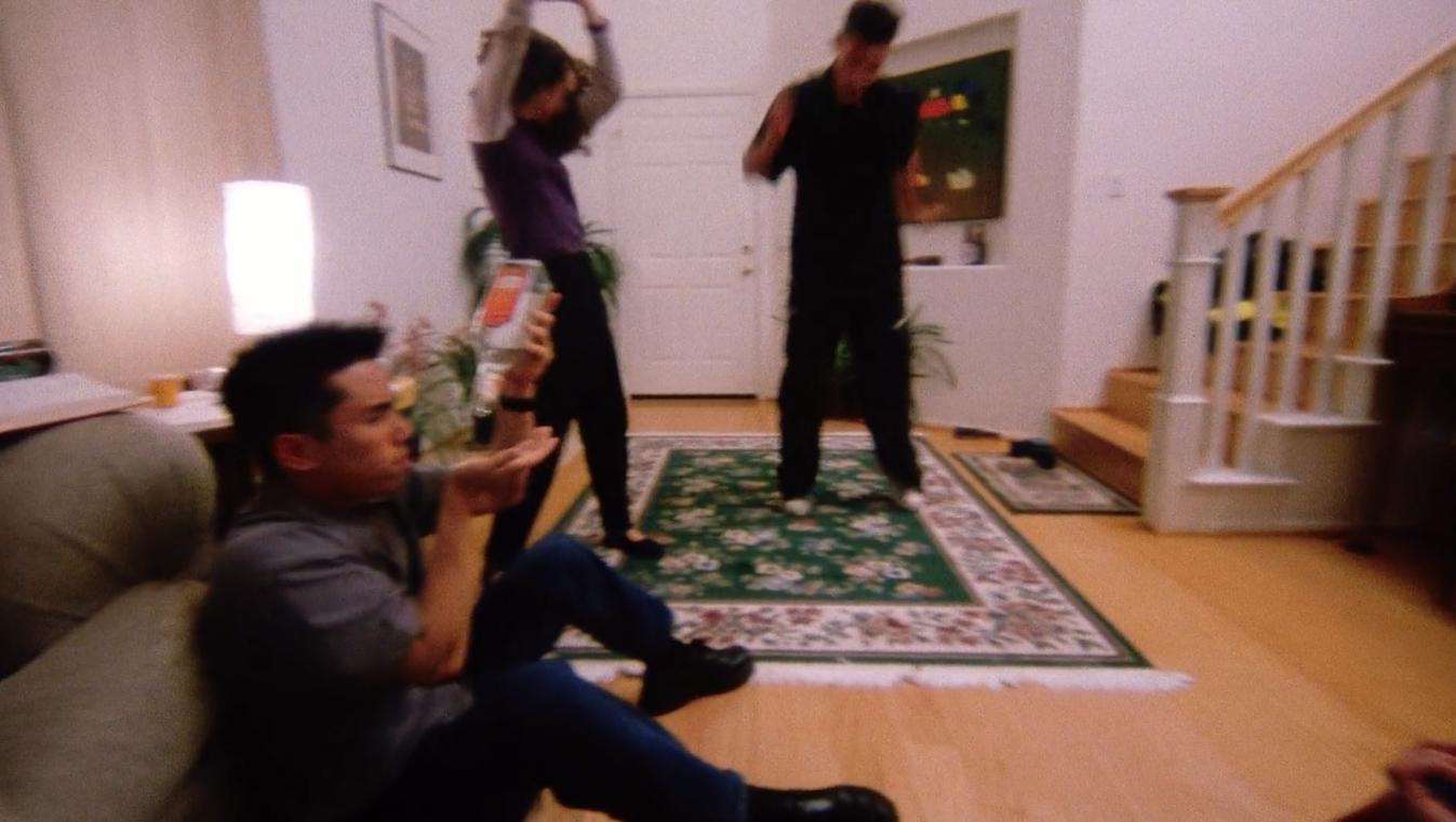 A blurry, wide shot of a portion of a house party. Two figures in the middle ground are dancing, while in the foreground, Ben, portrayed by Parry Shen, attempts to pour the last drops of an alcoholic beverage into his hand. 