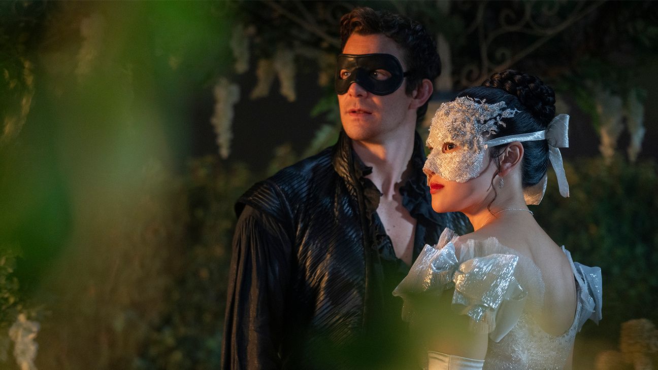 Benedict Bridgerton (Luke Thompson) and Sophie Baek (Yerin Ha) standing on a terrace outside a masquerade ball. Benedict is wearing all black, including a black mask, and Sophie is wearing a sparkling silver dress and mask. The two are standing close to each other and looking off to the left. Verica, Tom. Bridgerton. 2026.