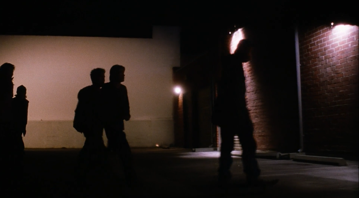 A dimly-lit shot of five shadowy figures. Two are on the far left, closer to the center are another two huddled together, and one is alone on the right side of the shot, in the foreground.