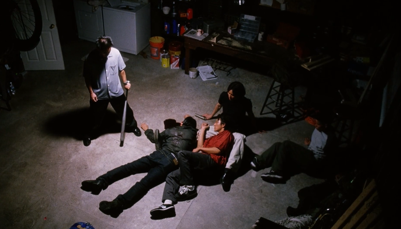The boys sprawl on the floor of a dimly-lit tool shed, staring at the prone form of Steve's lifeless body. Ben, the only one standing, holds a metal bat.