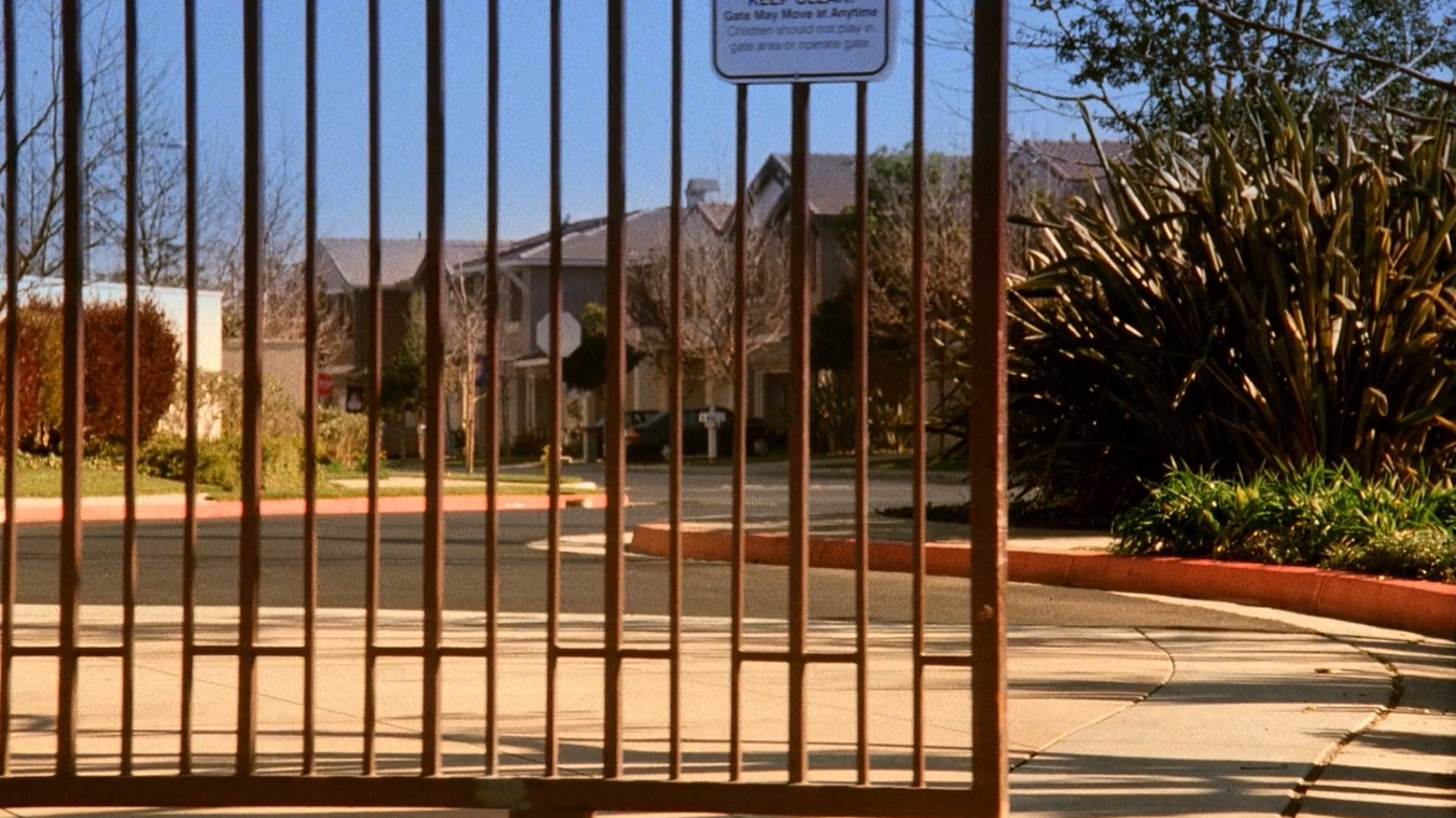 The view approaching the entrance to a suburban neighborhood, slightly obscured by a gate, which is sliding open for the viewer.