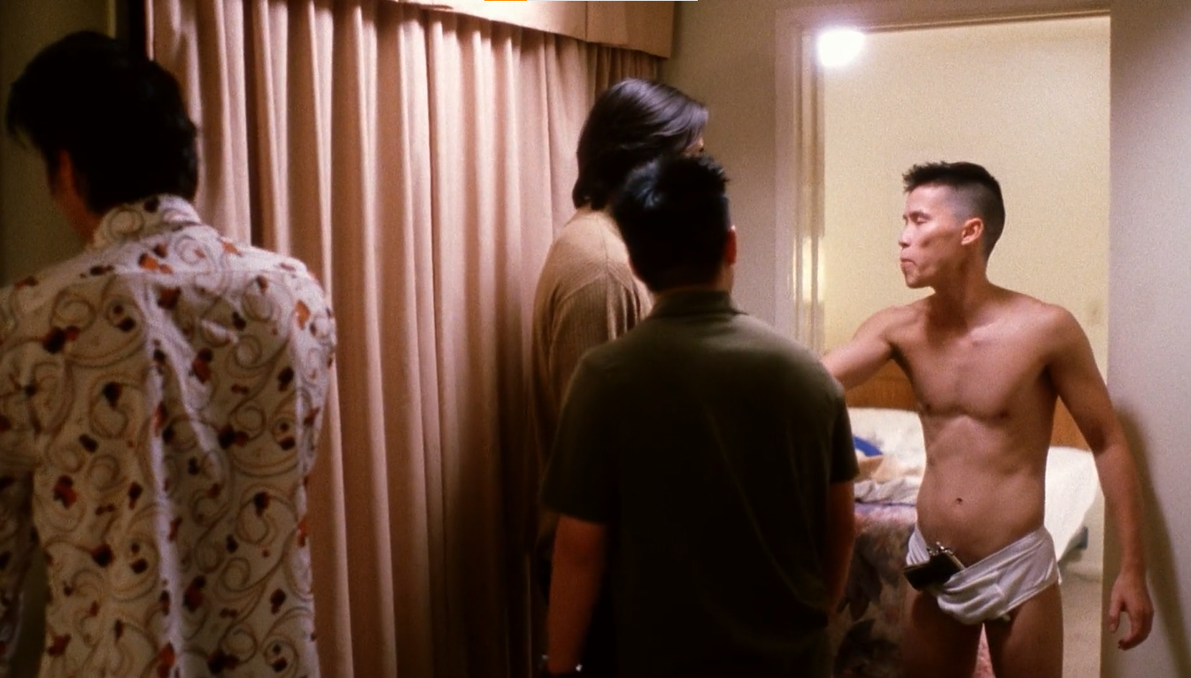 A wide shot of all the boys in the small foyer of a hotel room. All their heads are facing away from the viewer, save for Virgil on the right side of the frame. He stands in the doorway of a bedroom, wearing only underwear with a gun tucked into the crotch.