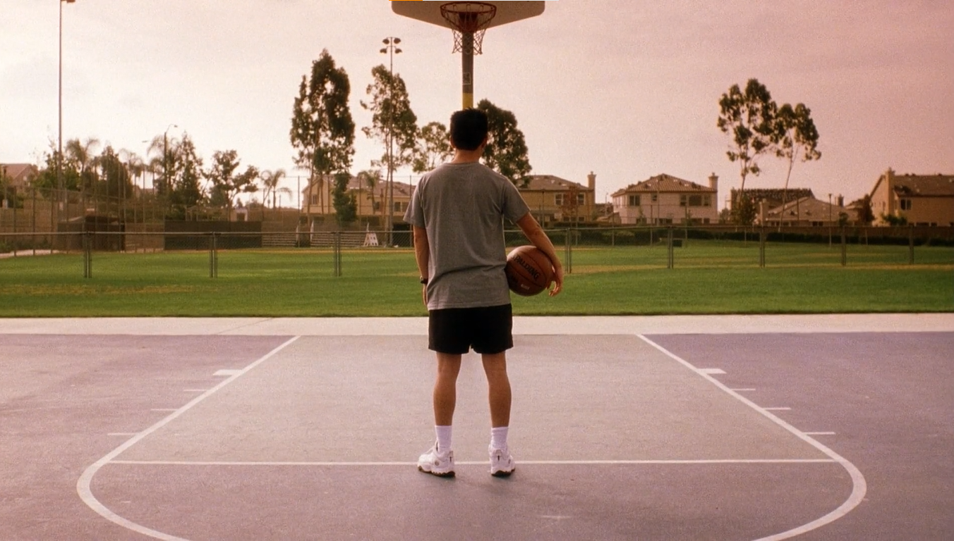 Ben, portrayed by Parry Shen, stands alone on a basketball court overlooking a manicured, green field. An array of suburban homes sit on the horizon line. 