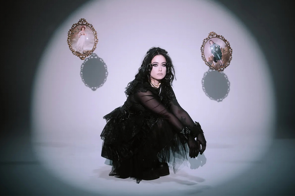 margo in an elaborate black gothic outfit crouches inside a bright spotlight, flanked by two floating, ornate framed portraits.