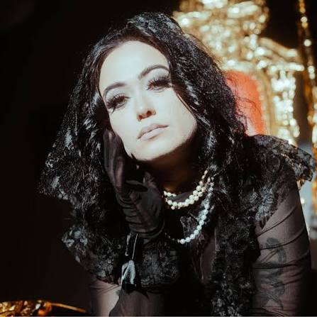 A close-up portrait of margø in a black gothic lace outfit, wearing pearls and leaning their chin on a gloved hand.