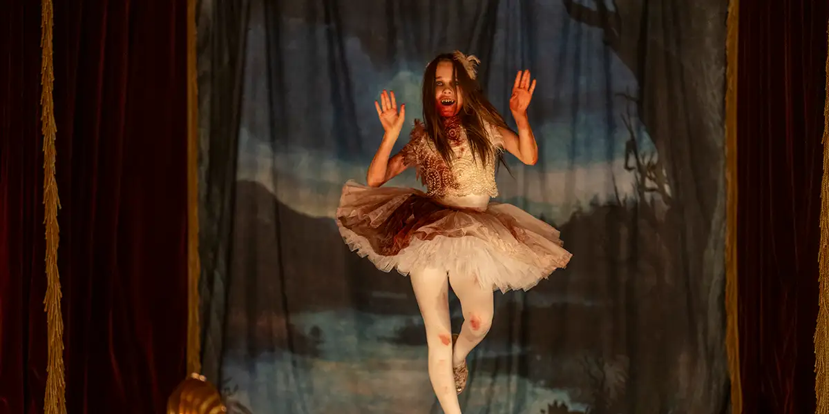 Alisha Weir as Abigail in Abigail performing a ballet dance on a stage. Her costume and mouth is covered in blood, with her sharp teeth on display.