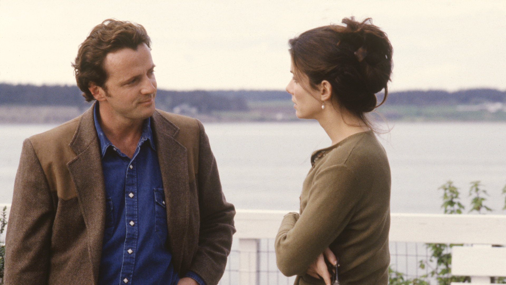 Practical Magic's Gary and Sally. They are standing outside with a water cove visible behind them as they face one another, having a conversation.