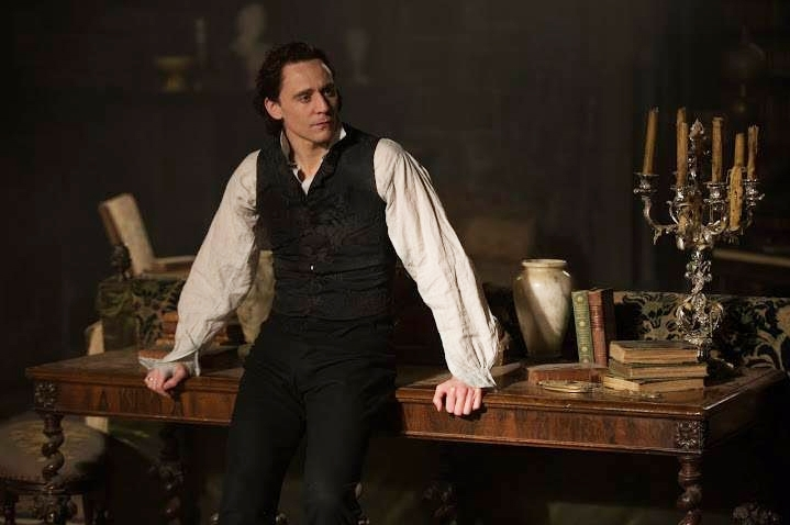 Thomas leans against the table of his workshop, watching with intent as Edith wanders around.