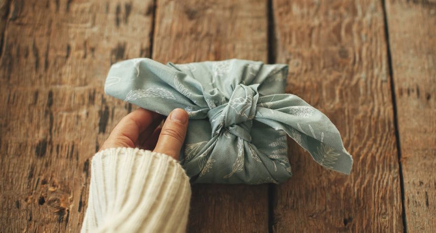 Furoshiki eco friendly present. Hand holding stylish gift wrapped in fabric on rustic wooden table. Happy holidays. Zero waste winter holidays, birthday or mothers day