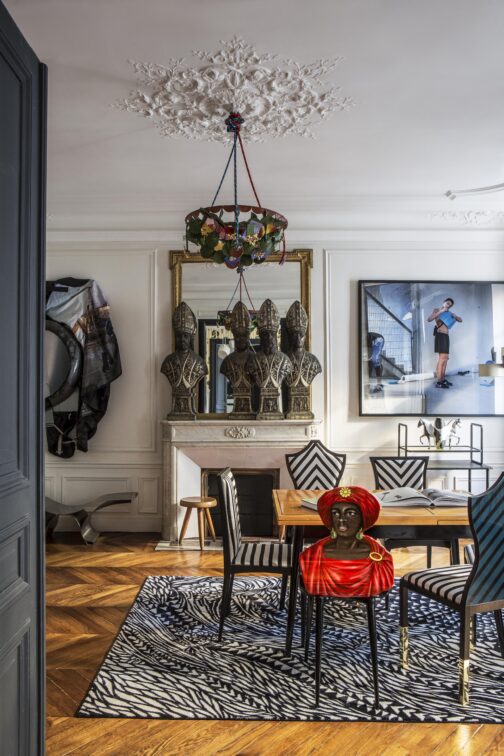 Chandelier: By Rodrigo Almeida, decorated with vintage jewels from Christian Lacroix Haute Couture. Sculptures (on mantlepiece): 4 Italian metal reliquary busts from the beginning of the 19th Century, from the gallery ‘Sans Prétentions’ in London. Stool: from Charlotte Perriand (from the 60s) Rug: ‘Wild Vibrations’ by Sacha Walckhoff for YO2 Design. Table: By Hans Bellmann (60s). Chair (red): ‘Moro’ by Piero Fornasetti. Chairs (stripes): from both Garouste and Bonnett and Christian Lacroix Maison. Upholstered in ‘Cabanon’ satin by Christian Lacroix Maison. Artwork (left): ‘Three or four times again’ by Mohamed Bourouissa from the Kamel Mennour gallery. Artwork (right): ‘R.I.P’ by Mohamed Bourouissa from the Kamel Mennour gallery. Bench: by Maria Pergay (70s). Table (back-right): By Jacques Adne Tray: ‘One Two Tray’ by Sacha Walckhoff for Edition van Treeck. Vase: ‘Equus’ vase by Sacha Walckhoff for Vista Alegre from the Maison Parisienne gallery