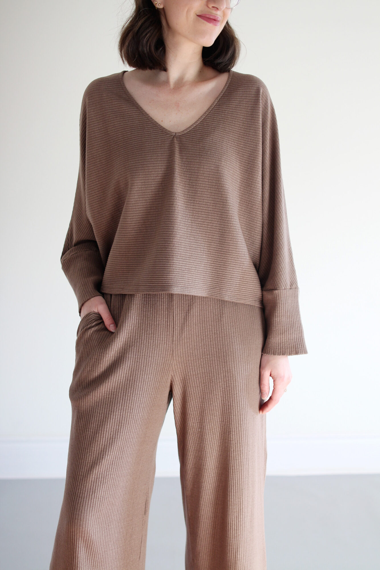 Woman wearing a cozy taupe cardigan over a matching minimalist lounge set in a taupe ribbed knit with fluffy slippers and clear framed glasses.