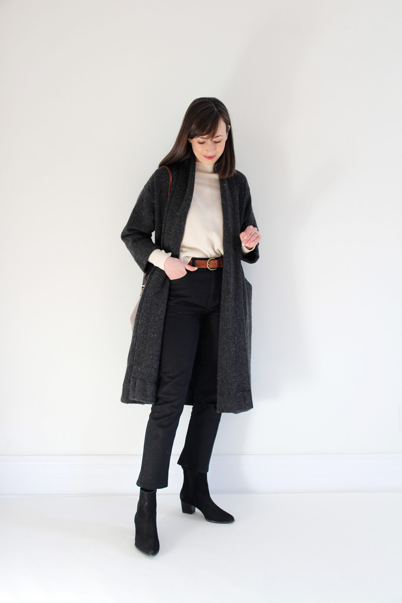 Woman wearing grey wool sweater coat, ivory mockneck sweater, brown leather belt, black jeans, black boots, and brown bag.