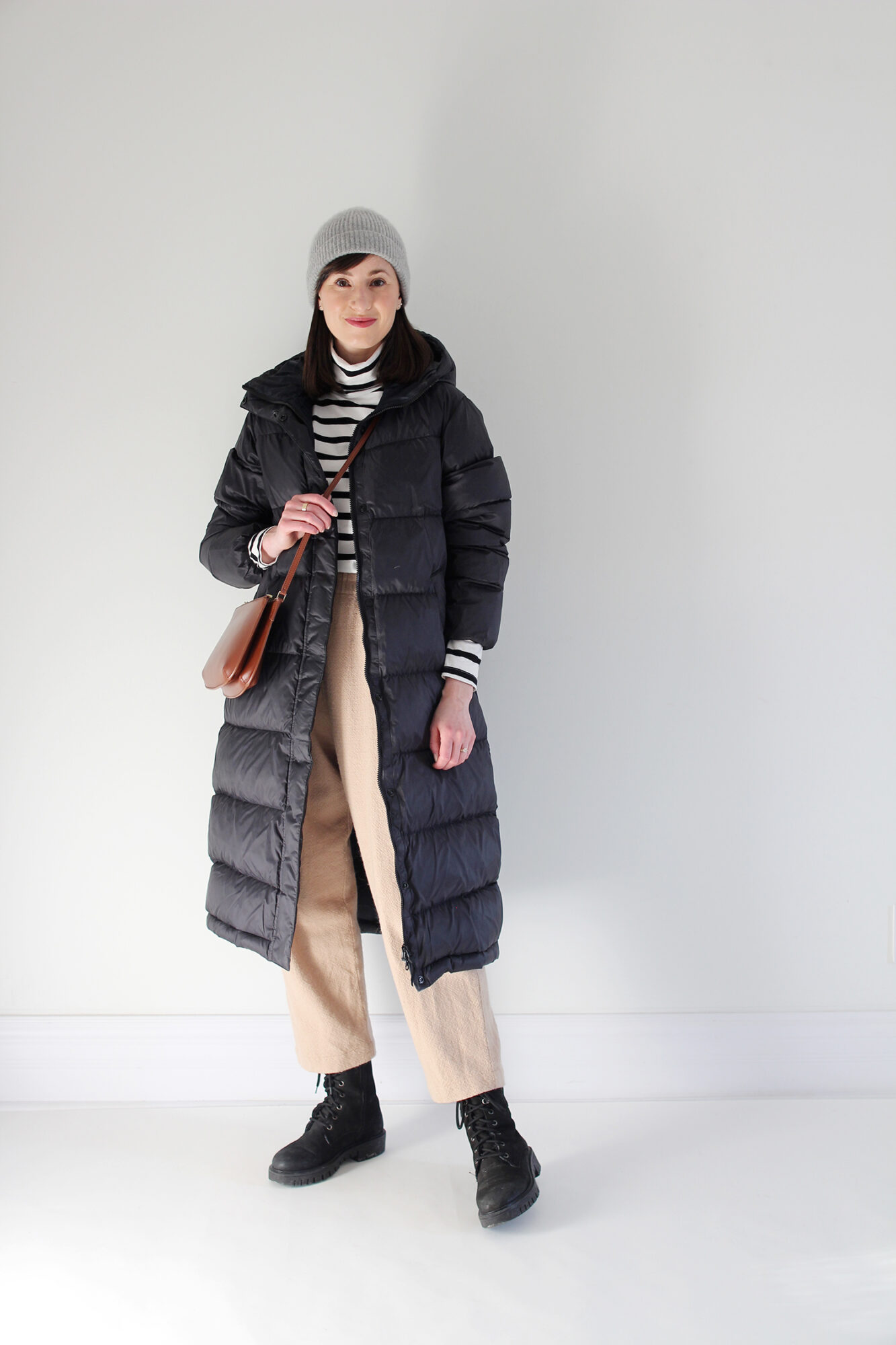 Woman wearing a long black puffer coat, striped turtleneck, tan wool trouser, black lace-up boot, grey beanie and brown leather bag.