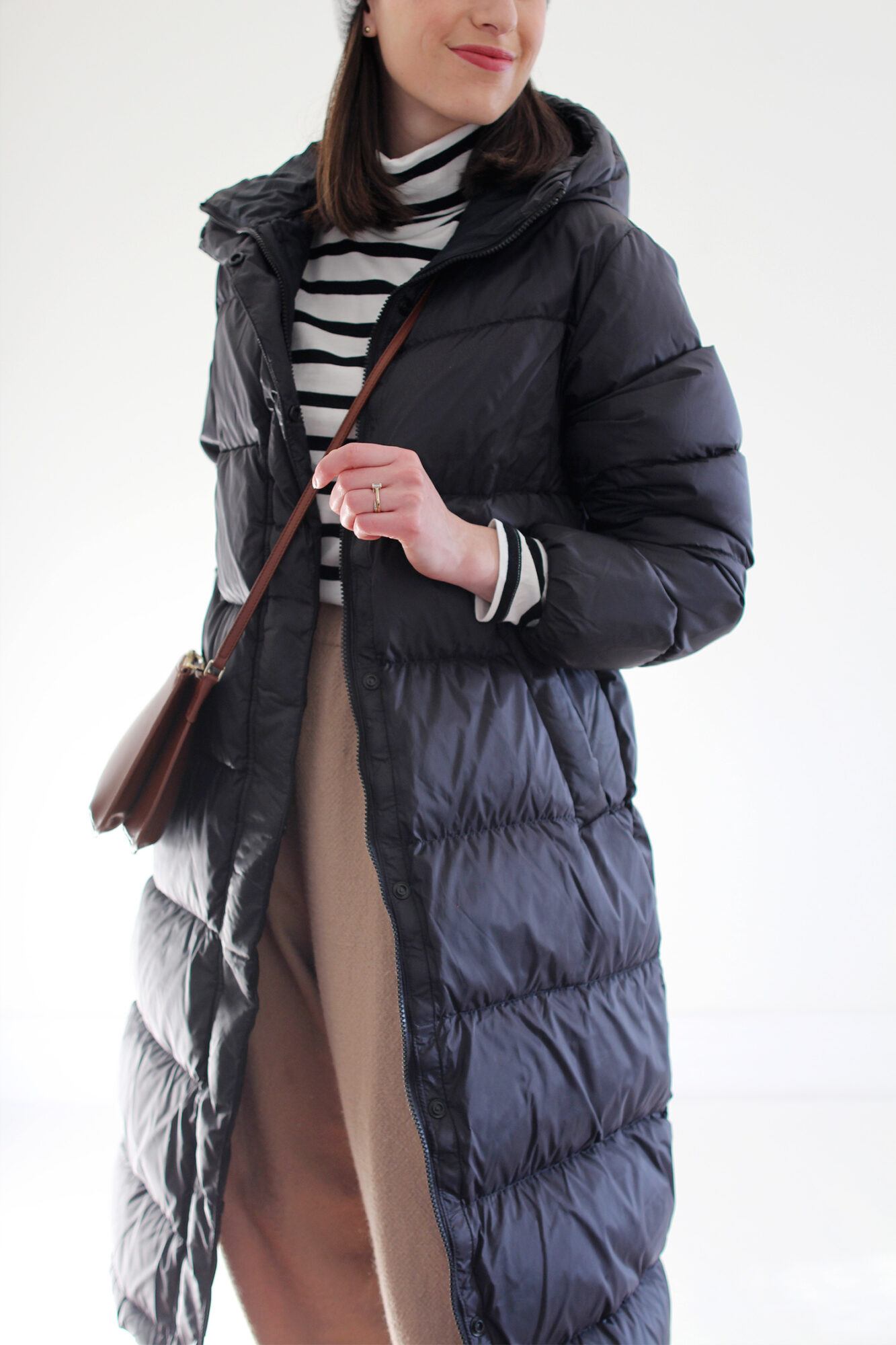 Woman wearing a long black puffer coat, striped turtleneck, tan wool trouser, black lace-up boot, grey beanie and brown leather bag.