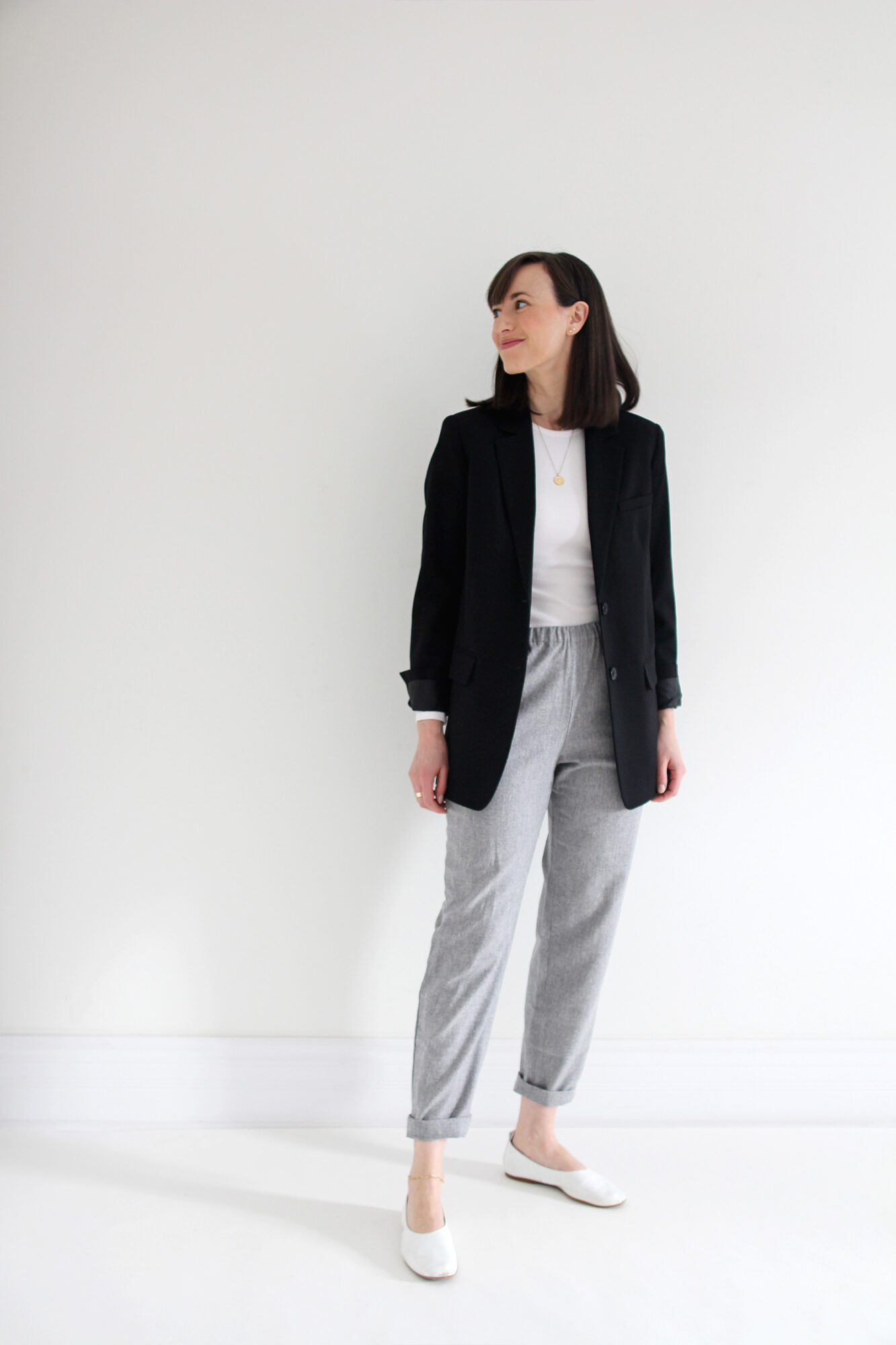 Woman wearing black blazer, white fitted t-shirt, grey joggers, white flats & a gold pendant necklace.