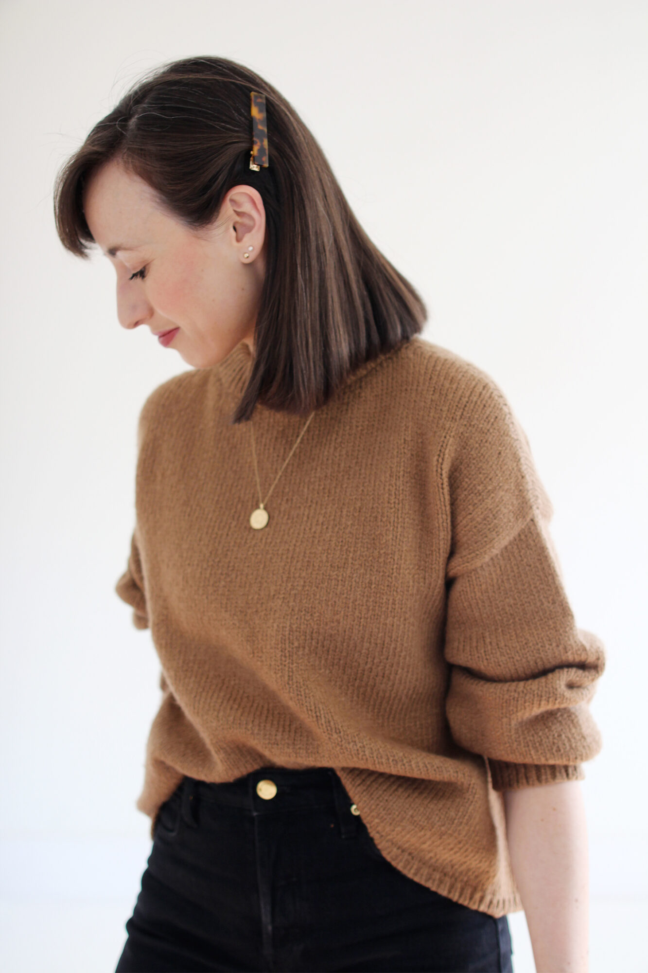 Woman wearing a relaxed alpaca knit sweater in camel, tucked into black jeans with gold jewelry and a tortoise barrette.