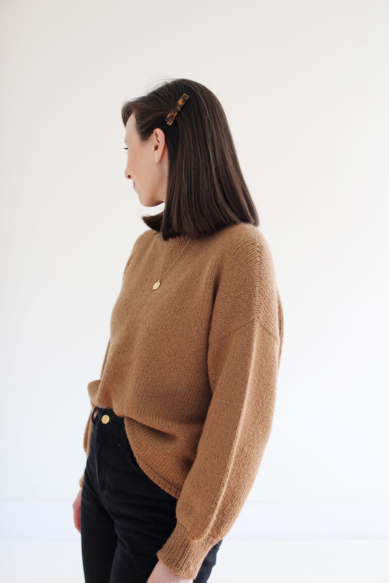 Woman wearing a relaxed alpaca knit sweater in camel, tucked into black jeans with gold jewelry and a tortoise barrette.