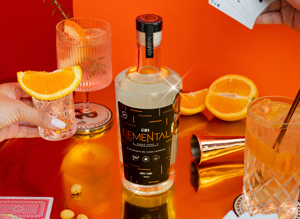 Botella de Gin Elemental sobre una mesa de vidrio con rodajas de naranja y tragos servidos de fondo.