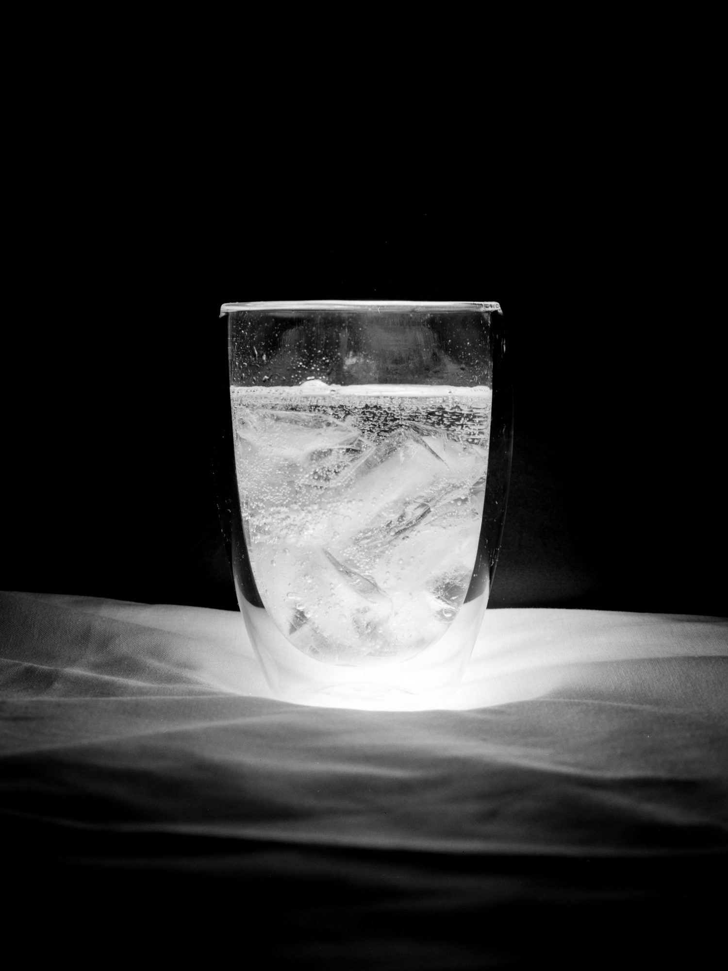 A black-and-white photograph of a glass of fizzy water with ice, brightly lit from below.