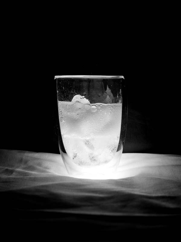A black-and-white photograph of a glass of fizzy water with ice, brightly lit from below.