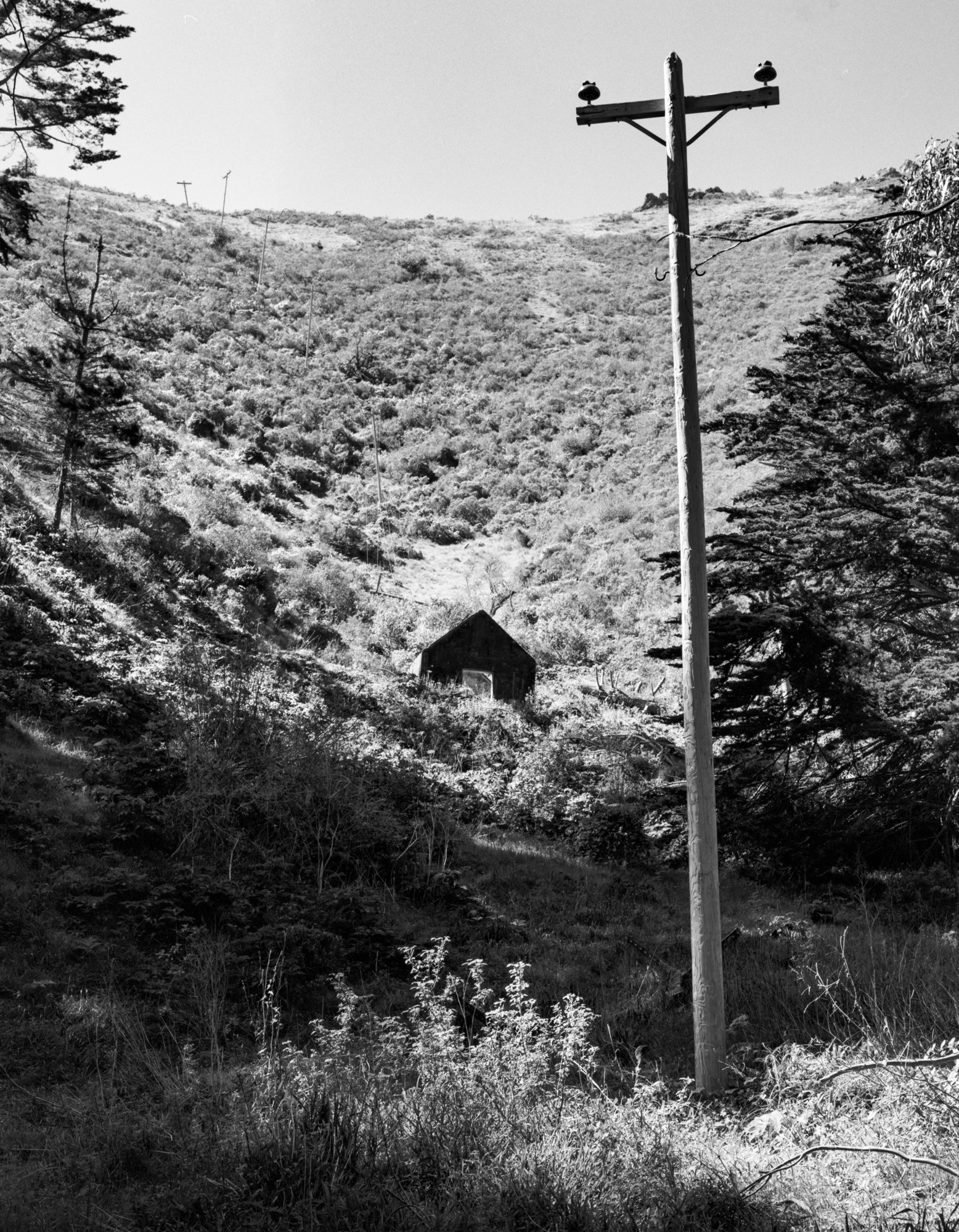 A black-and-white photograph of an abandoned building on the side of a steep hill with leaning and fallen power lines.