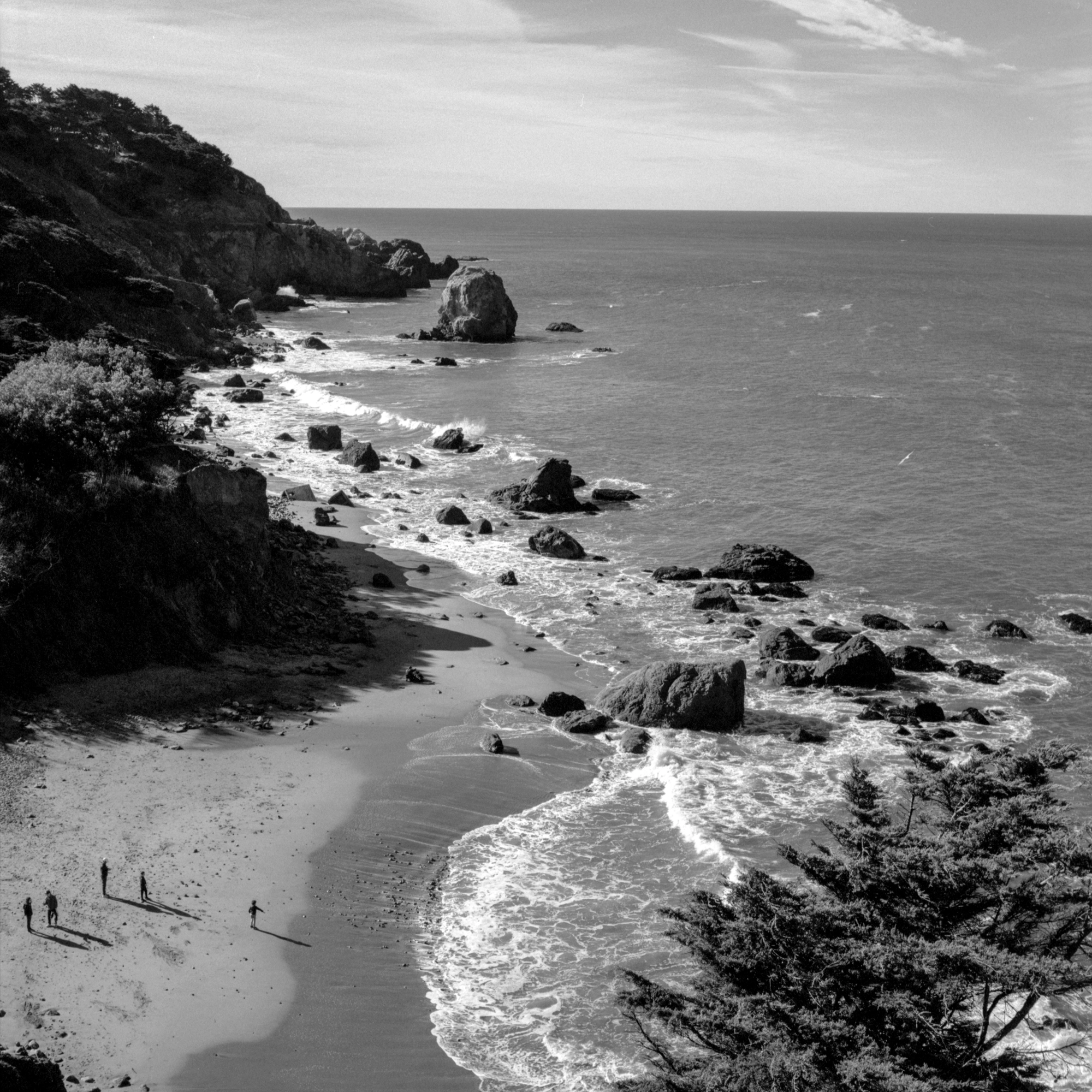 A black-and-white photograph of a rocky beach seen from above. In the bottom left corner, a child points toward the ocean.