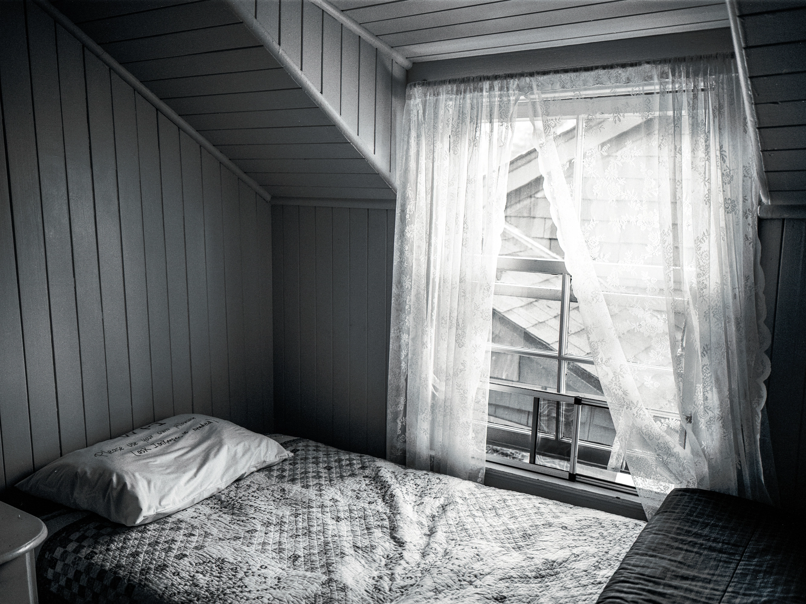 A black-and-white photograph of a bedroom in a historic inn with lacy curtains glowing from the setting sun.