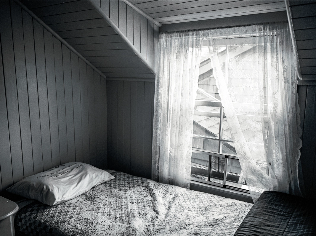 A black-and-white photograph of a bedroom in a historic inn with lacy curtains glowing from the setting sun.